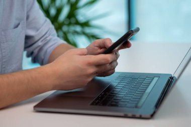 Ofiste akıllı telefonuyla yazı yazan bir adamın ellerini kapat. Sonra cep telefonunu bırakıp dizüstü bilgisayarda yazmaya başladı..