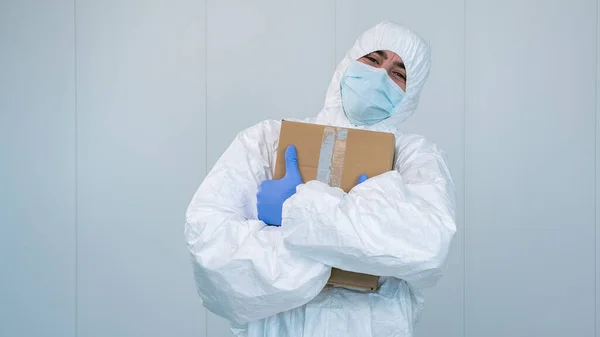 A happy health worker on protective suit hug a box of medical supplies with his arms in the hospital during pandemic caused by covid 19, coronavirus. The male nurse wears a surgical mask.