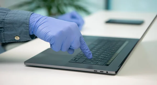 Person mit Handschuhen an den Händen, die die Enter-Taste auf seinem Laptop drückt. In der Nähe befindet sich ein Smartphone. Mitarbeiter in einem Büro während der Covid-19-Pandemie. — Stockbild Person Mit Handschuhen Den Händen Die Die Enter Taste Auf — Stockfoto