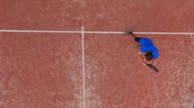 Açık hava sahasında raket oynayan genç bir adamın yukarıdan çekimi. Beyaz çizgilerin üzerinde yer alıyor..