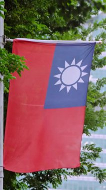 Arka planda yeşil bir ağaç ve şehir binası olan Tayvan bayrağının dikey yavaş hareketi doğayı kentsel gururla birleştiriyor. Çevresel ve kültürel temalar için ideal.