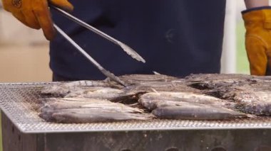 Fukuoka, Japonya-05 Ekim 2025: Ağır çekim bir aşçı Fukuoka, Japonya 'da dumanlı bir barbeküde balık pişiriyor, eldiven giyiyor ve açık hava gıda etkinliklerinde sıcak ışık altında hassasiyetle çalışıyor
