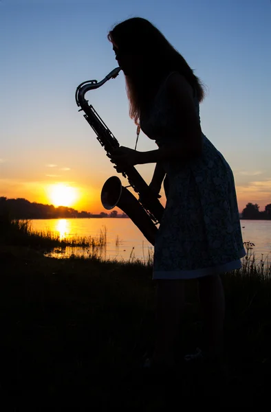 Silhouette eines Mädchens in einem Kleid in der Morgendämmerung in der Nähe des Flusses im Dorf spielt Saxophon vor blauem Himmel — Stockfoto