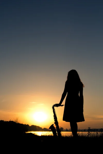 Silhouette of a woman in a dress on a background of a beautiful sun set and the sky saxophone on the grass and enjoy the view — Stock Photo, Image