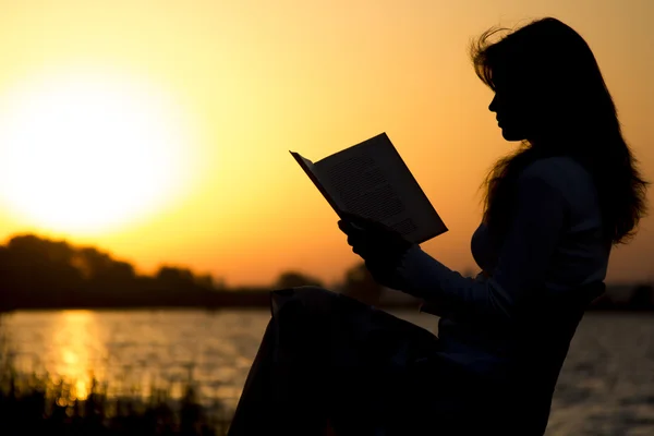 Silhouette of a young beautiful woman at dawn sitting on a folding chair and carefully staring at the open book — Stock Photo, Image