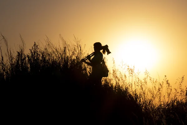 Silhouette einer Frau mit einer Kamera, die auf einem Stativ fixiert ist und bei Sonnenuntergang einen Berg hinaufgeht — Stockfoto