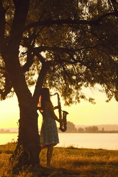 Silhouette portrait of beautiful young woman near the trunk of a tree at sunrise with sax — Stock Photo, Image