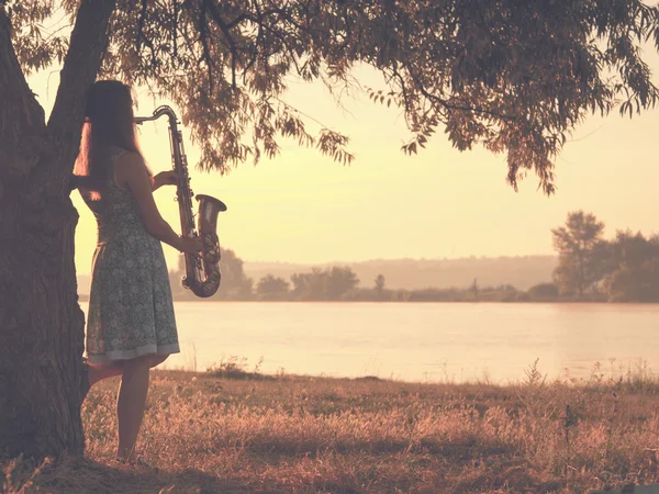 Ritratto vintage di una bella giovane donna in piedi vicino a un albero sulla riva del fiume con un sax — Foto Stock