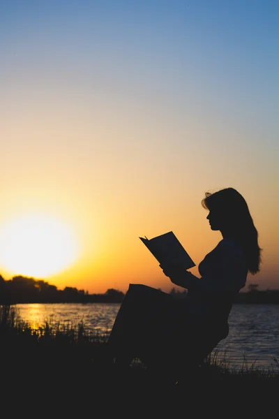 Contornos de una hermosa joven con un libro junto al río al amanecer del día — Foto de Stock