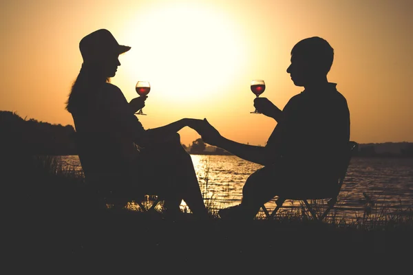 Obrysy mladých párů, uvolňující se na břehu řeky za úsvitu a sklenička vína na židlích — Stock fotografie