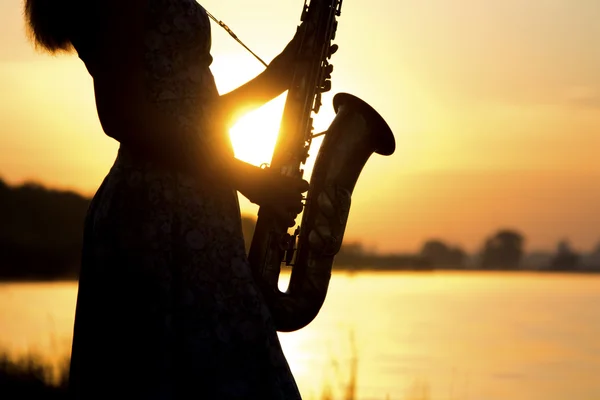 La silueta del saxofón en las manos de la mujer joven cerca del río al atardecer — Foto de Stock