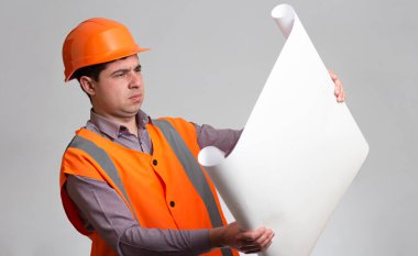 Sert şapka ve yansıtıcı yelek giyen genç inşaat işçisi, Gri stüdyo arka planında mühendis, kağıt üzerinde açılan projeden memnun değil.