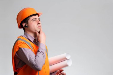 Sert şapka ve yansıtıcı yelek giyen genç inşaat işçisi gri stüdyo arka planında elleri çenesinde düşünüyor, ustabaşı bina projesini inceliyor.