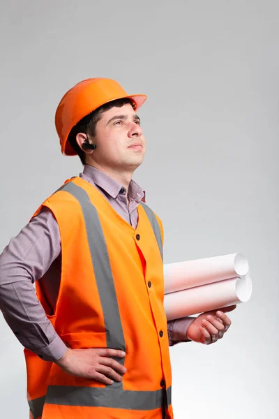 Young construction worker in hard hat thinking with scroll paper building project in hand on grey studio background, construction manager