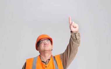 middle-aged builder in hard hat and reflective vest, protective glasses on grey studio background looking up and poke with finger