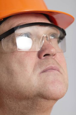 face of confident plump engineer, middle-aged carchitect in orange hardhat and protective glasses looking up on grey studio background