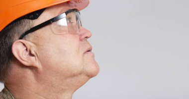 close-up face profile of thoughtful plump engineer in protective glasses, middle-aged construction worker in hard hat on grey studio background