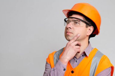 Kasketli ve yansıtıcı yelek giyen genç mühendis gri stüdyo arka planında el altında düşünerek endüstri kavramını inşa ediyor.