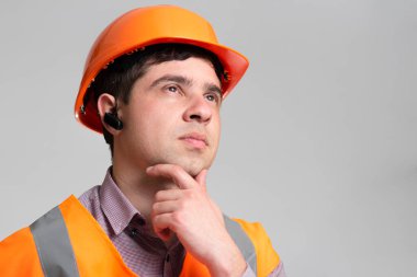 Sert şapkalı ve yansıtıcı yelekli genç mühendisin yüzü. Gri stüdyo arka planında çene altından düşünme, inşaat endüstrisi kavramı.