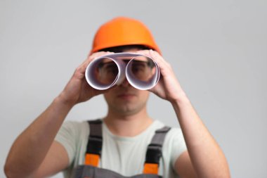 Gri stüdyo arka planında bir dürbün olarak taslak kullanarak sabit şapkalı ve tulumlu genç inşaat işçisi.