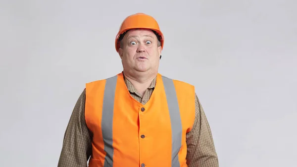 胖胖的建筑工人头戴安全帽 身穿灰色背景的反光背心 滑稽地混淆不清 不懂幽默的表情 — 图库照片