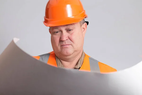 混淆不清 在结构上头戴硬礼帽的工人看着里面 手里拿着打开的纸 背景是灰色的工作室 工程师控制着 — 图库照片