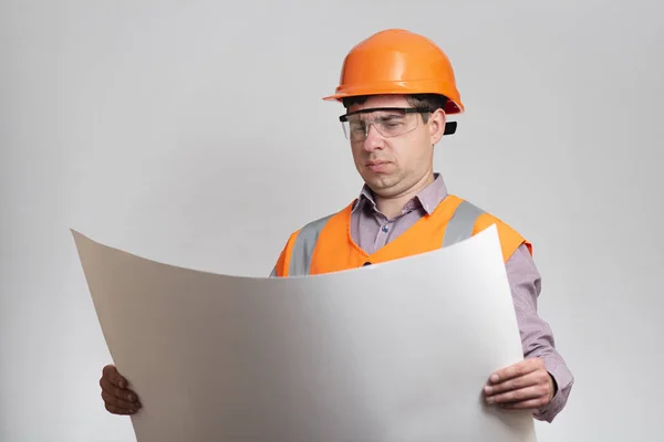 灰色のスタジオの背景に手をつないで開かれた紙プロジェクトとハード帽子の若い建設労働者 不満そうなエンジニア 顔の表情 — ストック写真