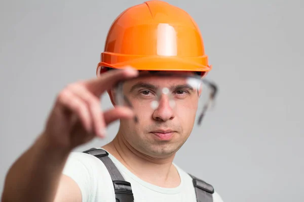 若い男の手の中にビルダーの制服の要素として保護眼鏡に焦点を当てヘルメットの建設労働者とグレーのスタジオの背景に全体 — ストック写真