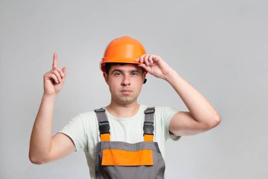 Şapkalı ve tulumlu kendine güvenen genç ustabaşı parmağını kaldırıyor, inşaat işçisi gri stüdyo geçmişini eşeliyor, inşaat sektöründe reklam yapıyor.