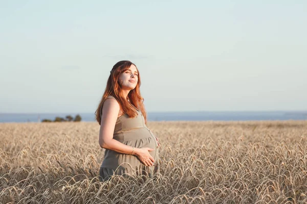 美丽的年轻孕妇在夕阳西下的时候走在成熟的麦田里 红头发的孕妇在大自然中放松地抚摸着她的肚子 快乐的怀孕观念 — 图库照片