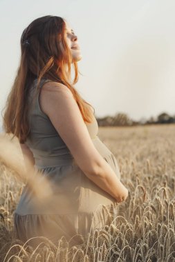 Gün batımında buğday tarlasında genç ve güzel bir kadın. Minnettarlıkla yukarı bakıyor. Kızıl saçlı, hamile bir anne.