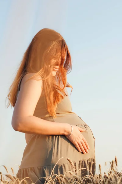 美丽的年轻孕妇在日落时走在麦田上 红头发的孕妇在大自然中放松地用手抚摩她的肚子 快乐的怀孕观念 — 图库照片