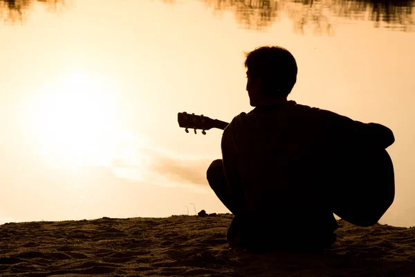 Silhouette eines jungen Mannes, der Gitarre spielt und im Sand sitzt — Stockfoto