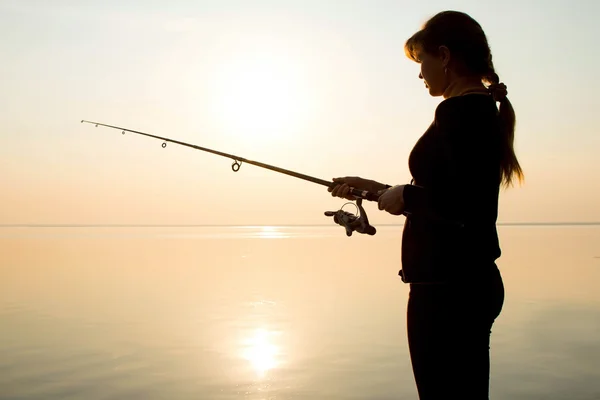 Silhouette di una giovane ragazza che pesca al tramonto vicino al mare — Foto Stock