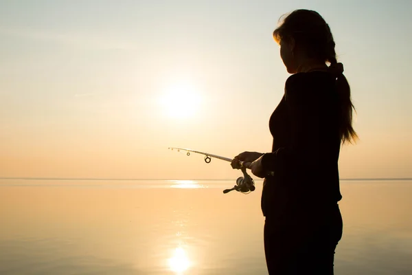 Silhouet van een jong meisje vissen bij zonsondergang in de buurt van de zee — Stockfoto