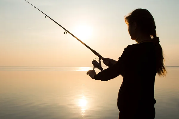 Silueta de una joven pescando al atardecer cerca del mar — Foto de Stock