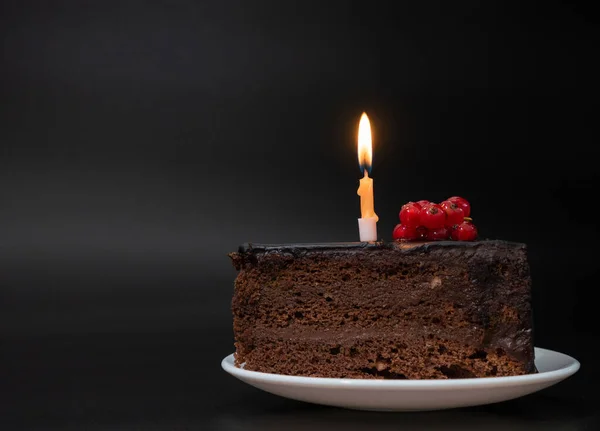 Kawałek czekoladowego tortu urodzinowego z jedną palącą się świecą na białym talerzu na głębokim czarnym tle. Urodziny, świętowanie, rocznica. Zbliżenie, kopiowanie miejsca na tekst. — Zdjęcie stockowe