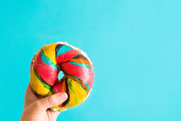 Bagel colorido com queijo e polvilhas na mão sobre fundo azul — Fotografia de Stock