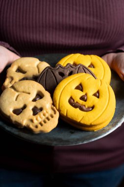 Hands holding plate with Halloween cookies shaped like pumpkins, skulls and bats on wooden table