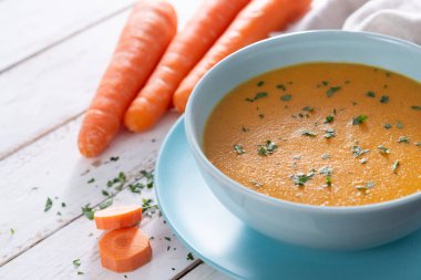 Ev yapımı kremalı havuç çorbası açık mavi bir kasede servis edilir, taze maydanozla süslenir ve kırsal beyaz ahşap bir masaya yerleştirilir..