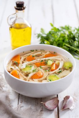 Beyaz kasede ev yapımı tavuk çorbası, beyaz ahşap masada havuç ve kereviz.