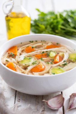 Beyaz kasede ev yapımı tavuk çorbası, beyaz ahşap masada havuç ve kereviz.
