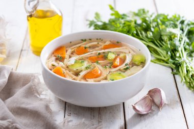 Beyaz kasede ev yapımı tavuk çorbası, beyaz ahşap masada havuç ve kereviz.