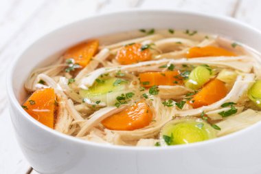 Beyaz kasede ev yapımı tavuk çorbası, beyaz ahşap masada havuç ve kereviz. Kapat.