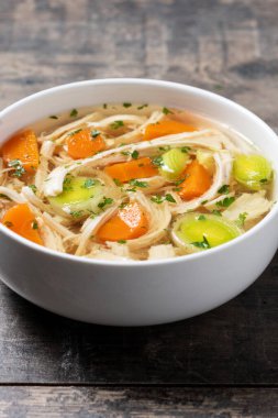 Beyaz kasede ev yapımı tavuk çorbası, beyaz ahşap masada havuç ve kereviz.