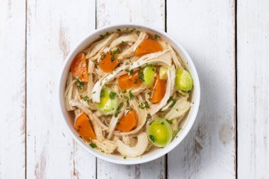 Beyaz kasede ev yapımı tavuk çorbası, beyaz ahşap masada havuç ve kereviz. Üst görünüm