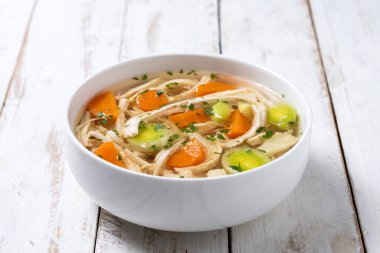 Beyaz kasede ev yapımı tavuk çorbası, beyaz ahşap masada havuç ve kereviz.