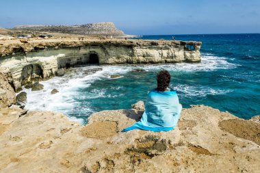 Girl sits on a ledge of rock above the sea at Cape Greco . Cypru clipart