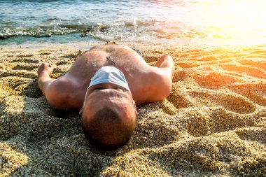 Tıbbi maskeli bir adam deniz kenarındaki bir tatil köyünde güneşleniyor.