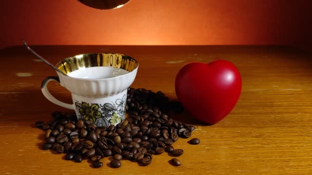 Кофе наливают в чашку, стоящую среди кофейных зерен рядом с игрушечным красным сердцем — стоковое видео